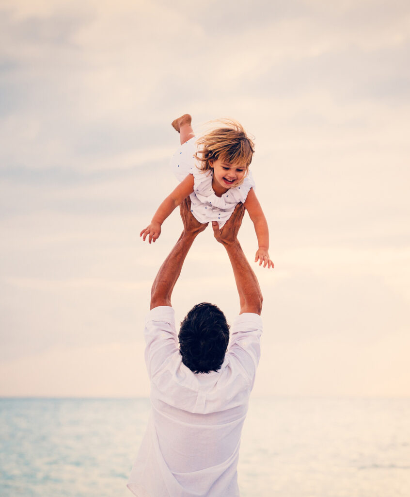 Padre aupa en el aire a su hija jugando junto a la playa en la puesta del sol